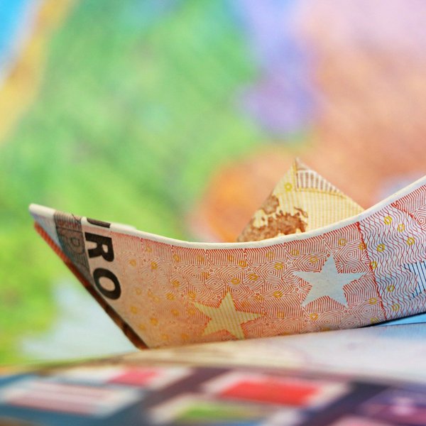 Das Bild zeigt ein aus einem Geldschein gebasteltes Schiff vor einem bunten Hintergrund.